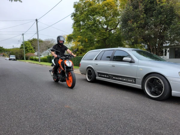 KTM Duke rider in helmet on suburban street with DD Roadworthies Ford Falcon wagon parked nearby.