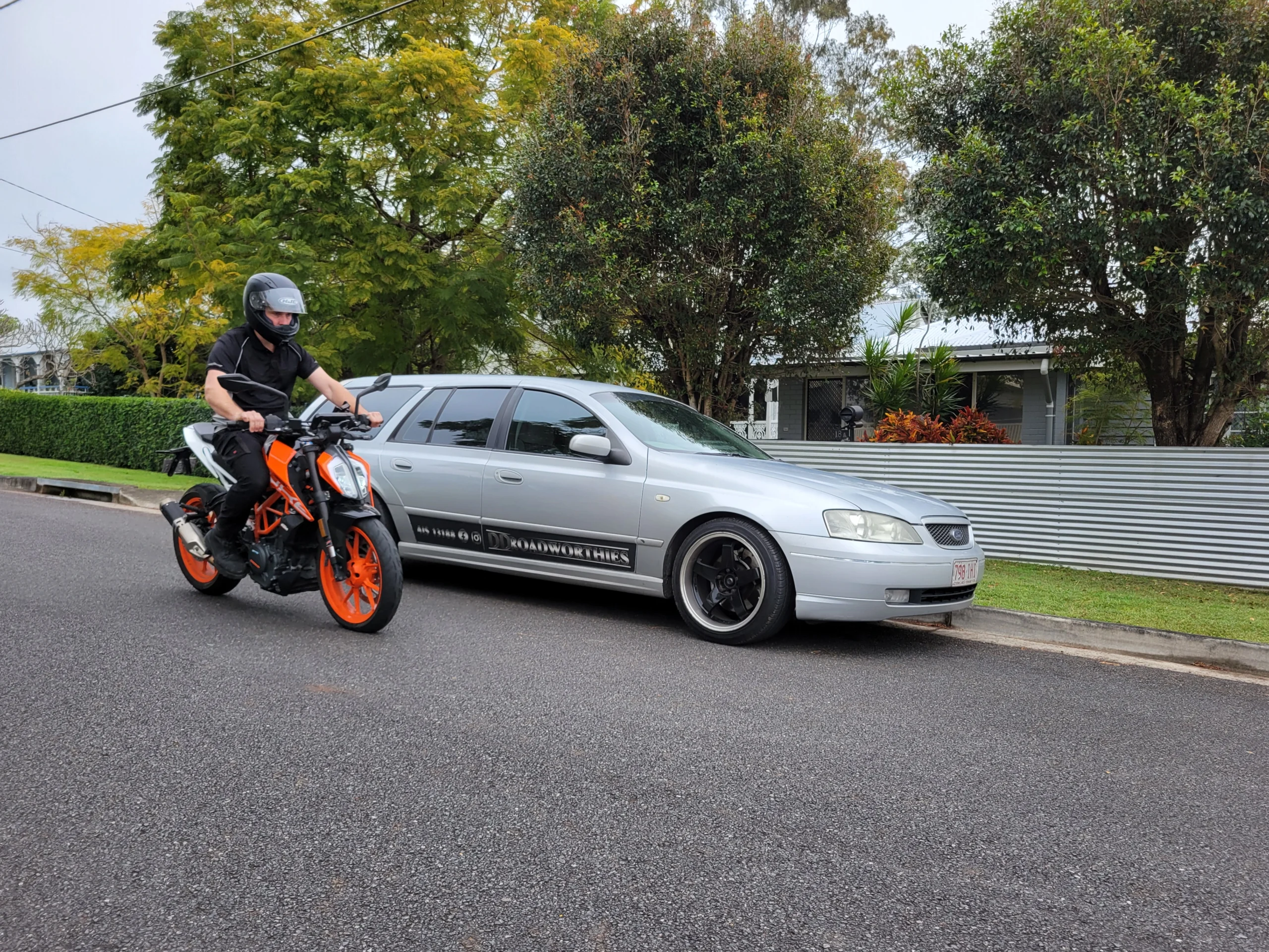 Rider on KTM Duke motorbike near DD Roadworthies Ford Falcon wagon during inspection service.