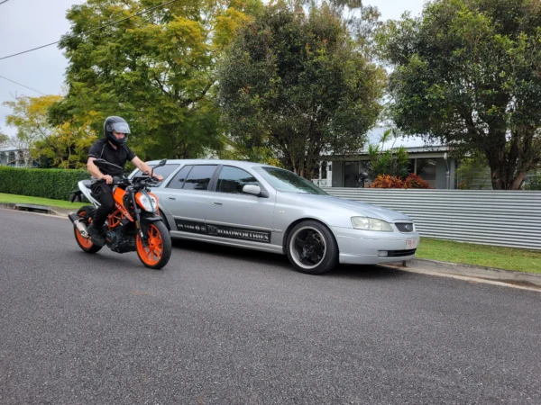 Rider on KTM Duke motorbike near DD Roadworthies Ford Falcon wagon during inspection service.