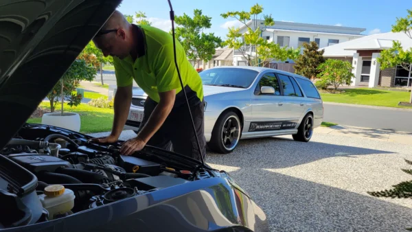 Mechanic inspecting Subaru BRZ engine with Ford Falcon wagon in background during roadworthy check.