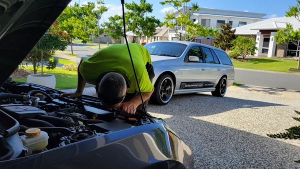 Mechanic leaning into Subaru BRZ engine bay for detailed roadworthy inspection at driveway.