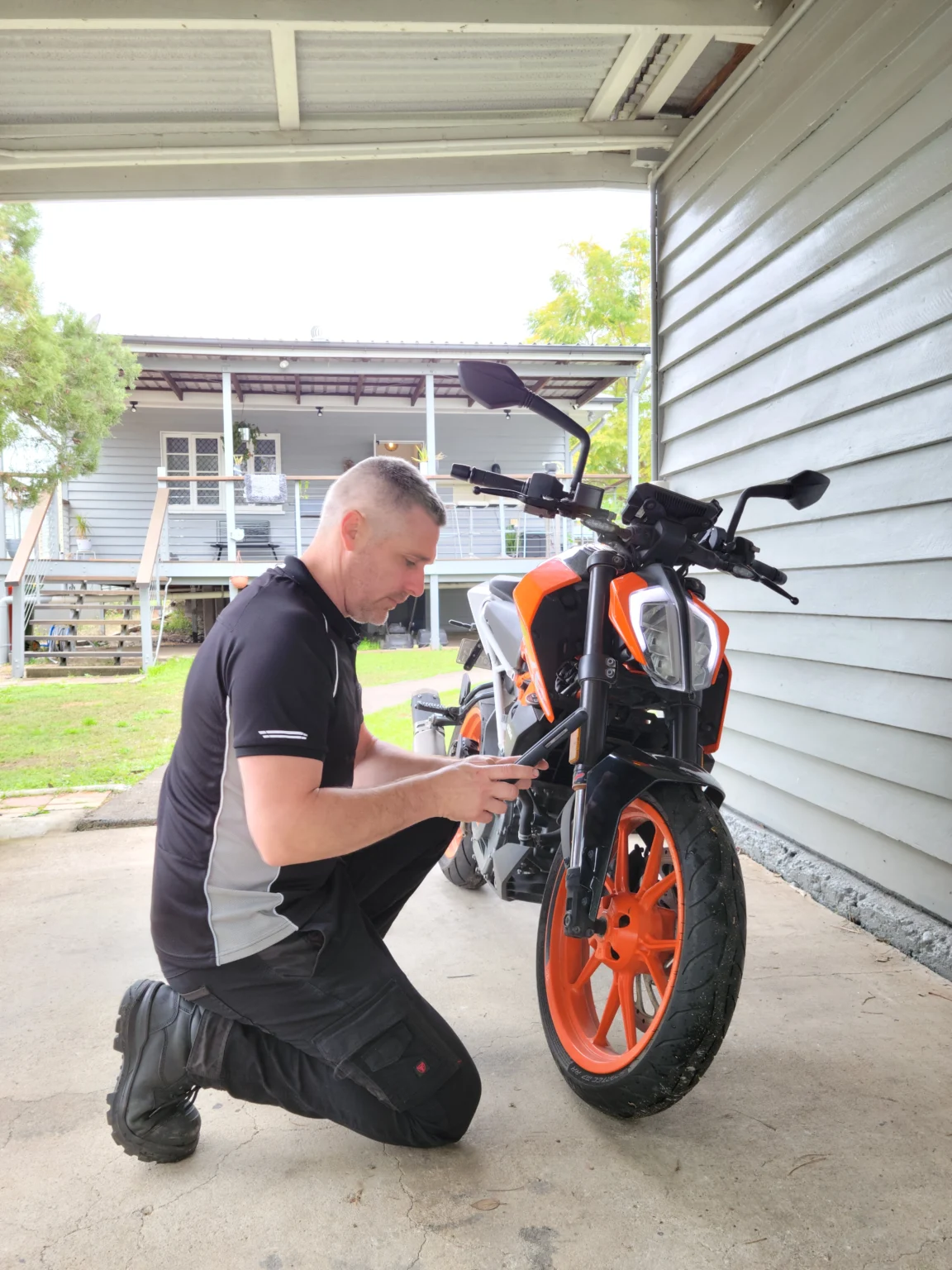 Mechanic kneeling beside KTM Duke motorbike performing safety inspection with tablet.